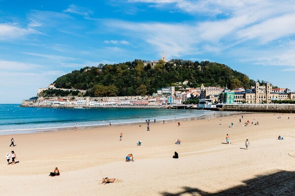 A beach with people sunbathing and swimming, a hill with buildings, and a clear sky.