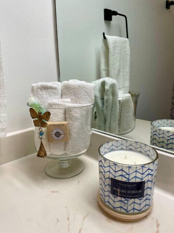 Un baño con un jabonera de vidrio con una pastilla de jabón, un candelabro de vidrio con una vela y un perchero con una toalla blanca.