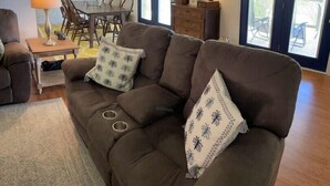 A living room with a brown sofa, two patterned throw pillows, a wooden coffee table, a dining table with chairs, a lamp, and a TV on a wooden cabinet. - Kerr Contentment   Kerr Lake/Buggs Island waterfront, an amazing view (Clarksville)