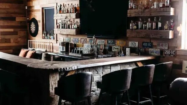 Uma área de bar com balcão de pedra, banquetas pretas, um alvo de dardos e prateleiras com garrafas de bebidas.