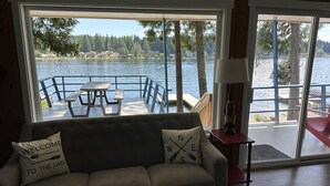 A cozy room with a large window overlooking a lake, a wooden deck, and a sofa with a 'Welcome to the Lake' pillow. - Charming Lakefront Cabin on Mason Lake with Pebble Beach and Private Dock (Grapeview)