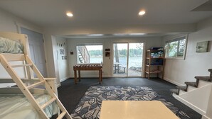 A room with a bunk bed, a foosball table, a desk, and a view of a lake through large windows. - Charming Lakefront Cabin on Mason Lake with Pebble Beach and Private Dock (Grapeview)