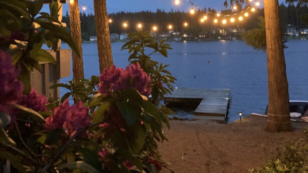 A lakeside patio with string lights, a wooden deck, and a bench. - Charming Lakefront Cabin on Mason Lake with Pebble Beach and Private Dock (Grapeview)