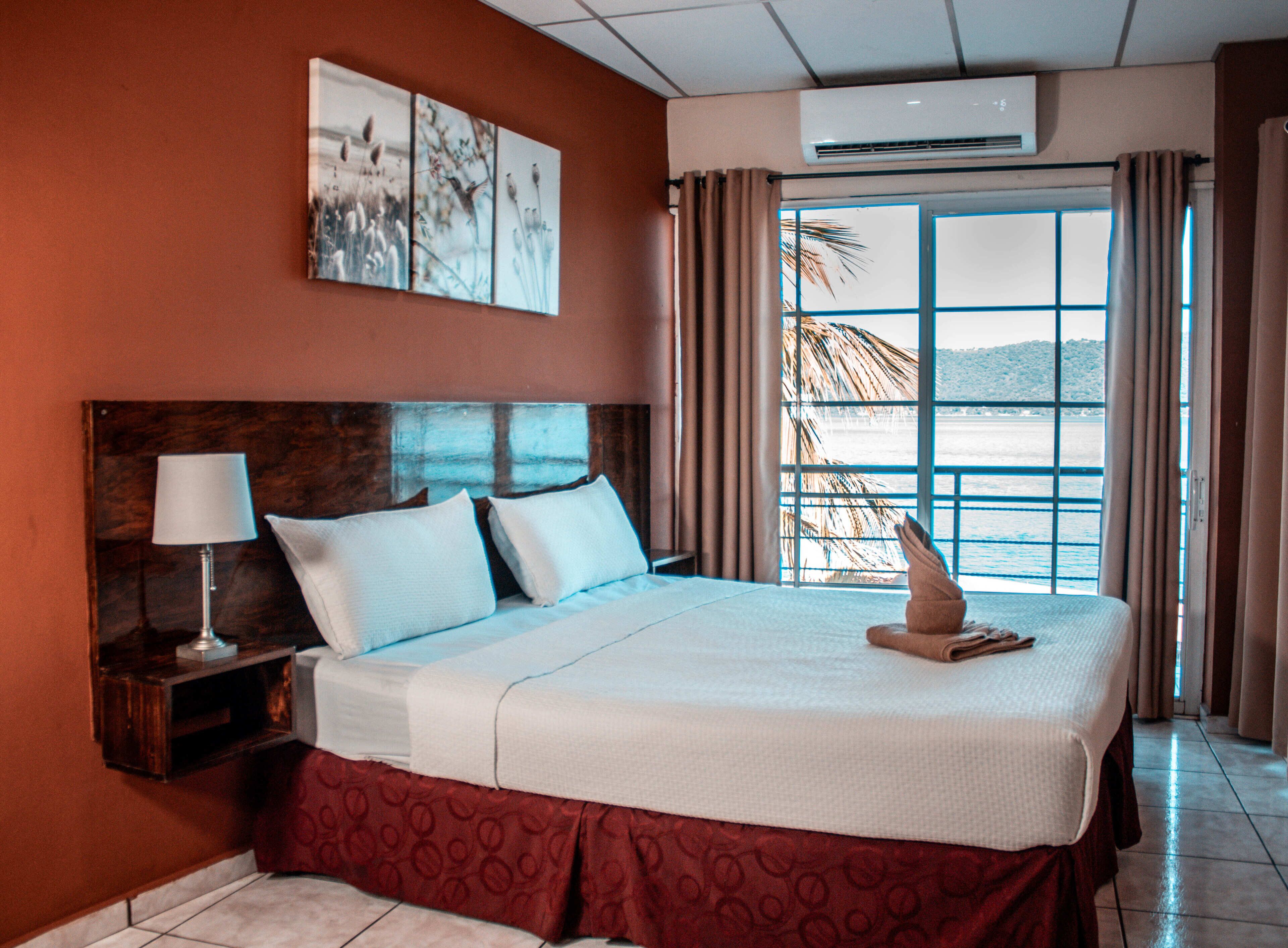 Habitación de hotel con una cama grande, una mesita de noche con lámpara, una ventana con cortinas y una obra de arte en la pared.