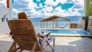 A person relaxing on a wooden lounge chair by a pool with a view of the sea and a wooden structure in the background.
