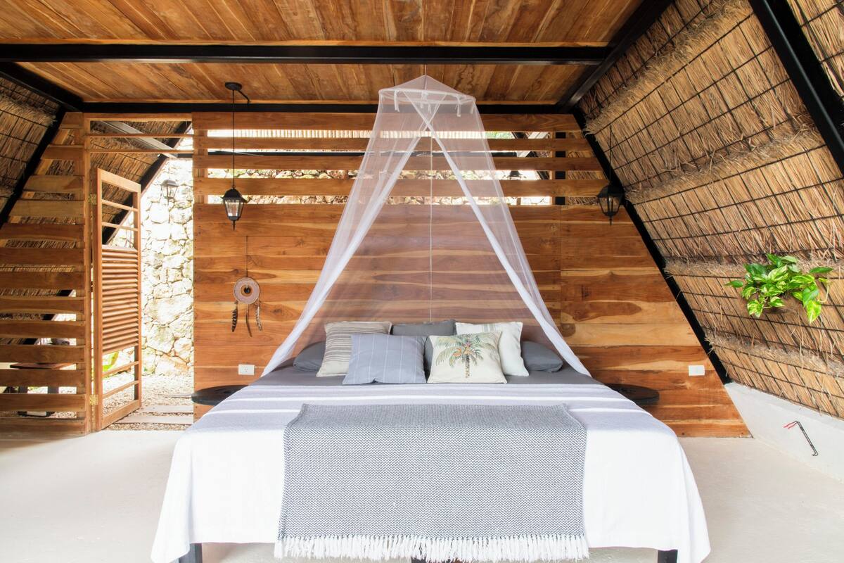 Une pièce en bois avec un lit recouvert d’une moustiquaire, un mur en pierre et une plante.