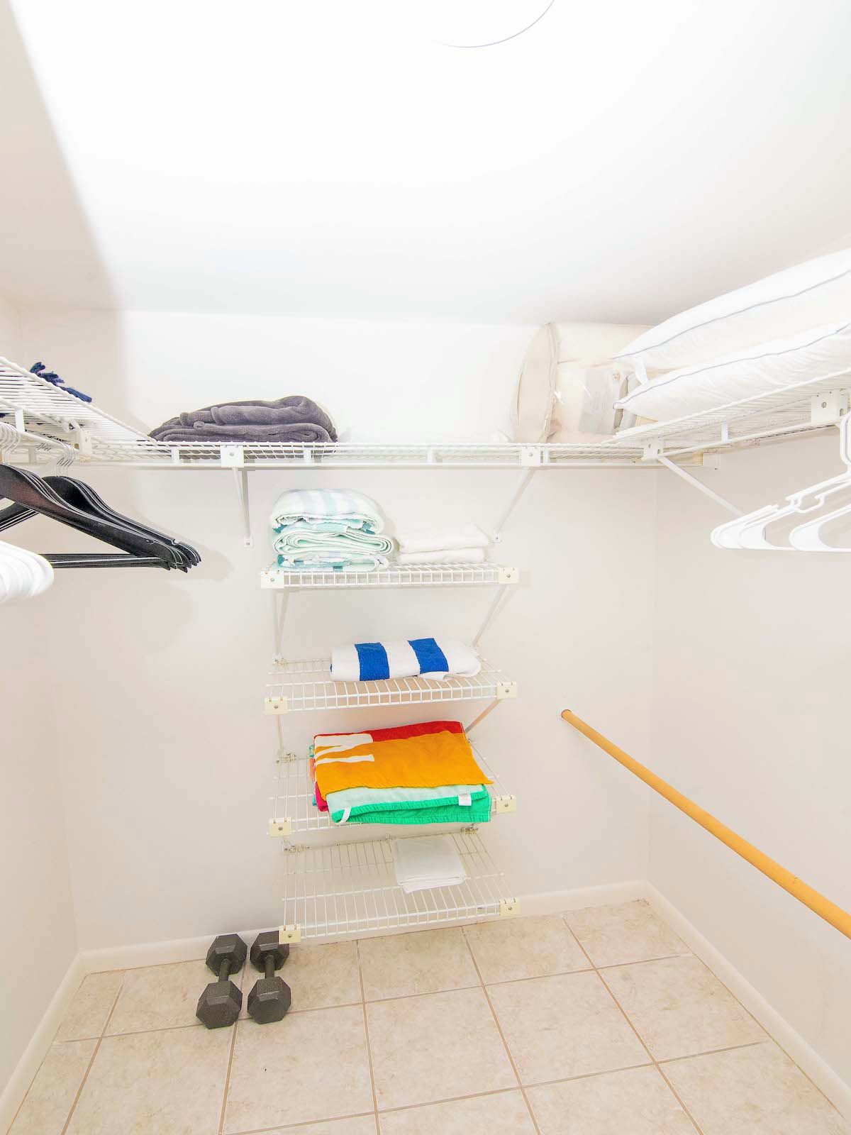 A storage closet with shelves for folded clothes, a hanging rack for hats, and a pair of dumbbells on the floor.