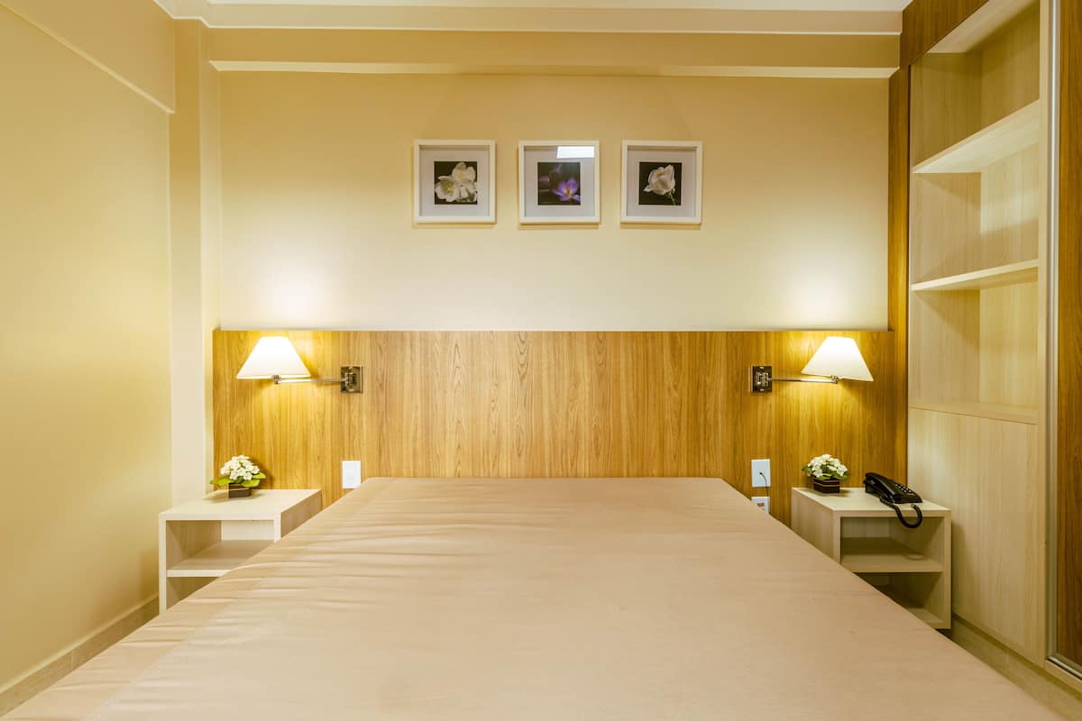 Habitación de hotel con cabecera de madera, dos mesitas de noche con lámparas y tres cuadros en la pared.