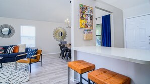 Una sala de estar moderna con un sofá, sillas, una mesa de centro y un comedor. Hay un tablón de anuncios en la pared y una puerta con cortinas.