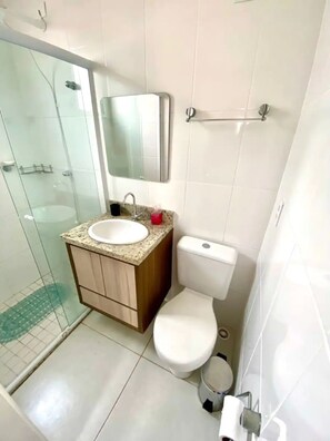 A bathroom with a glass shower enclosure, a sink with a mirror above it, and a toilet. - Apto novo e arejado com uma ampla sacada (Bertioga)