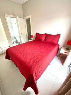 A bedroom with a large bed, a nightstand, a red lamp, and a bathroom visible through an open door. - Apto novo e arejado com uma ampla sacada (Bertioga)
