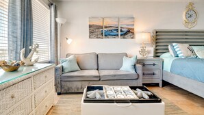 Ein gemütliches Schlafzimmer mit grauem Sofa, einem Bett mit blauer Bettwäsche, einem Nachttisch mit Lampe und einer Wanduhr.