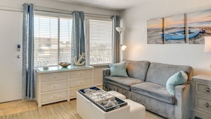 Ein gemütliches Wohnzimmer mit grauem Sofa, einem weißen Couchtisch und einer Körbschublade. Ein großes Fenster mit blauen Vorhängen lässt natürliches Licht herein.