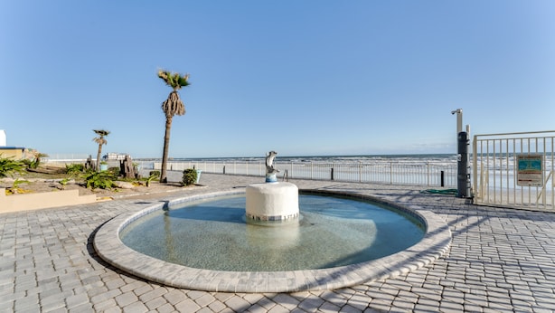 Ein runder Springbrunnen mit einer Statue in der Mitte, umgeben von einem gepflasterten Weg und Palmen.