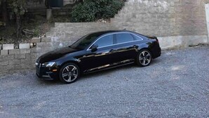 Une voiture noire était garée sur une allée de gravier devant une maison dotée d’un mur en pierre et d’une terrasse en bois.