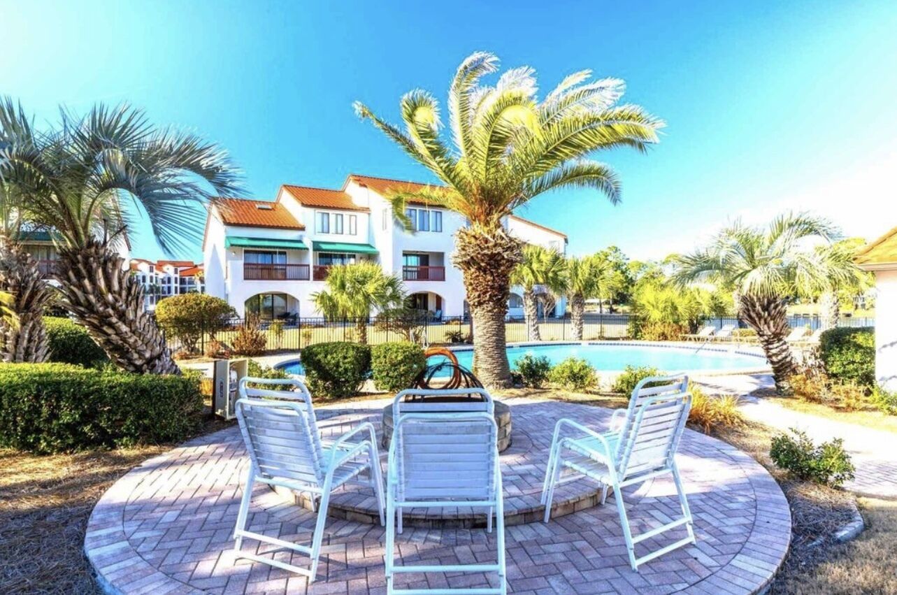 A poolside area with lounge chairs, a palm tree, and a building with balconies in the background.