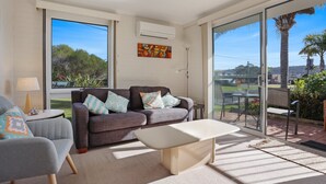A modern living room with a grey sofa, a white coffee table, and a large window offering a view of palm trees and a distant landscape. - Calendo Apartmnts - Unit 1: Ground Floor (Merimbula)