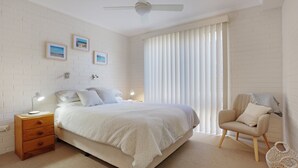A bedroom with a bed, a nightstand, a chair, and a window with blinds. - Calendo Apartmnts - Unit 1: Ground Floor (Merimbula)