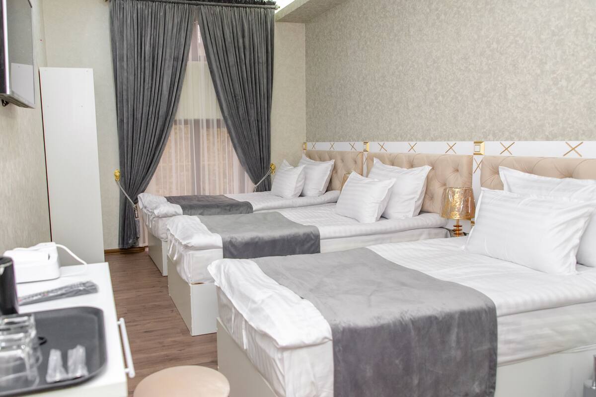 Habitación de hotel con tres camas, un escritorio con computadora y lámpara, y una ventana con cortinas.