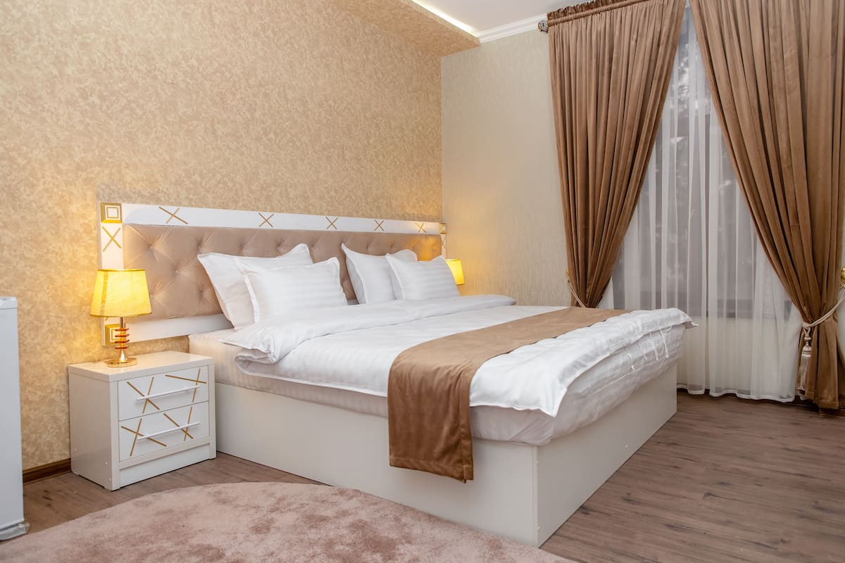 Un dormitorio moderno con cabecera capitonada, ropa de cama blanca, una mesita de noche con lámpara y un ventanal con cortinas.