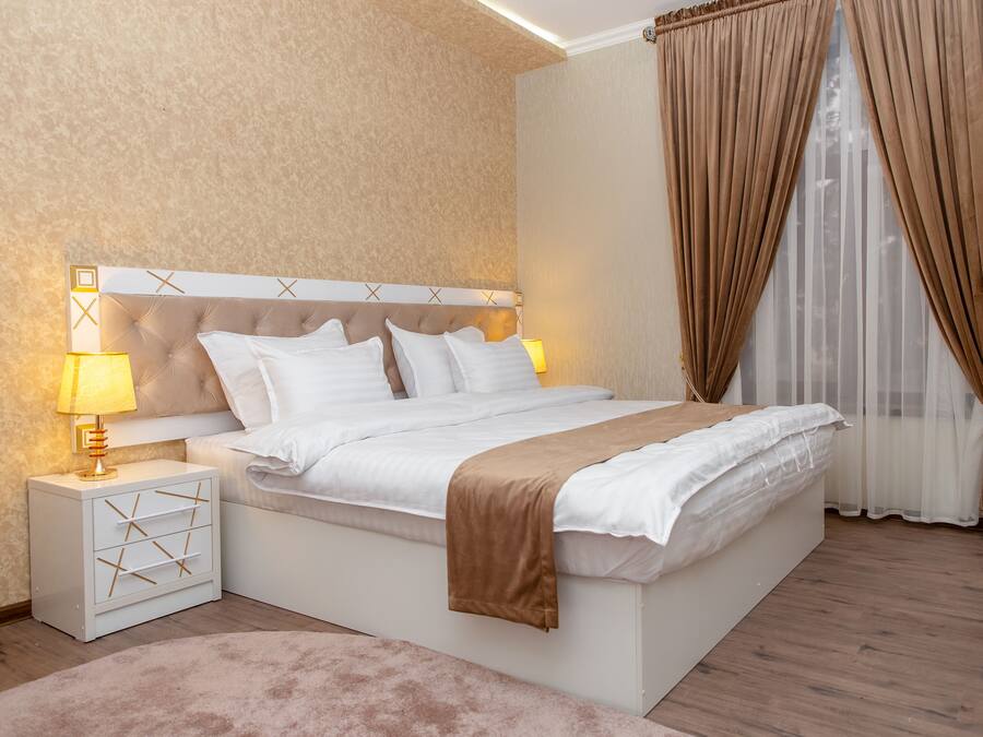 A modern bedroom with a tufted headboard, white bedding, a bedside table with a lamp, and a large window with curtains.