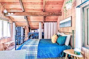 Une chambre dans une cabane en bois, avec un lit, une petite table, une plante suspendue et une fenêtre.