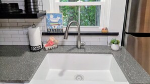 Une cuisine avec un évier en acier inoxydable, une fenêtre donnant sur les arbres, une étagère avec du liquide vaisselle et un panier.