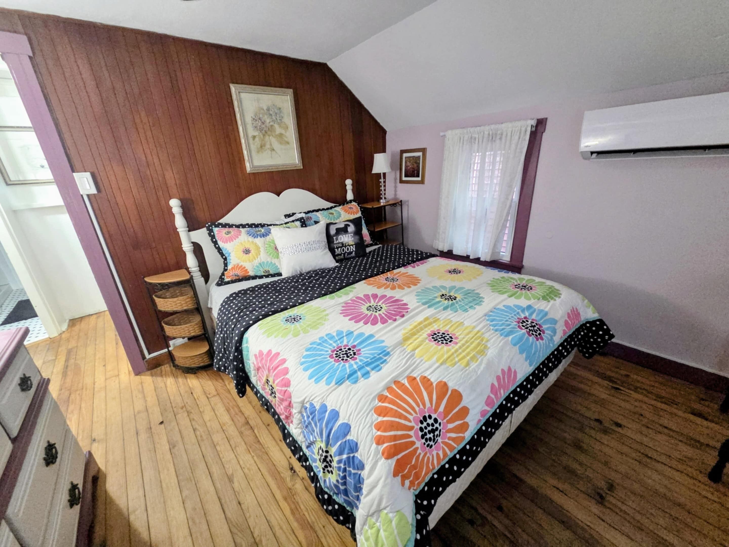 Ein Schlafzimmer mit Holzboden, einem Bett mit farbenfroher Blumenbettdecke, einem weißen Kopfteil und einer Wand-Klimaanlage.