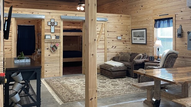 A rustic cabin interior with a wooden ceiling, a dining table, a sofa, and a small kitchen area with a sink and cabinets.