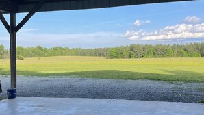A covered outdoor area with a concrete floor, a metal roof, and a view of a green field and trees in the distance.