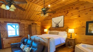 A log cabin bedroom with a bed, a sofa, a ceiling fan, a window, and a bathroom with a bathtub. - Romantic Cabin with Jacuzzi Tub. Ride-in Access to Wolf Pen Gap ATV trails  (Mena)