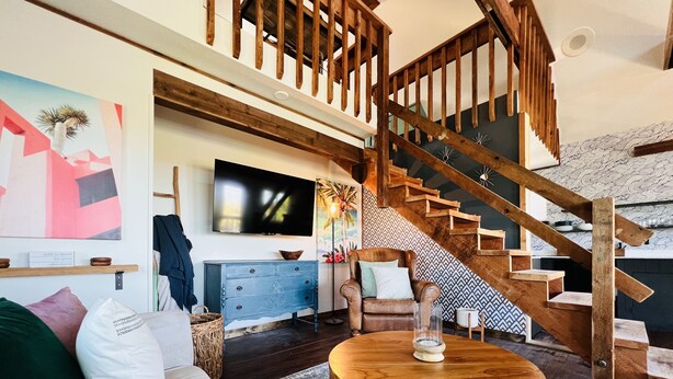 A modern living room with a wooden staircase, a flat-screen TV, a blue cabinet, and a round wooden coffee table. - The Pink Palm - Waterfront - Boat & RV’s Welcome! (Crystal River)