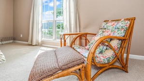 Une pièce avec une grande fenêtre, un fauteuil en osier agrémenté d’un coussin à motifs floraux et un plaid rayé posé sur le sol.