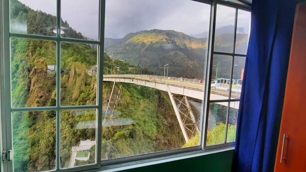Ein Zimmer mit großem Fenster und Blick auf eine Brücke über einem Tal, im Hintergrund Berge.