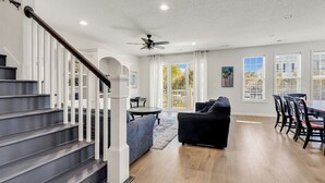 A spacious living area with a staircase, a ceiling fan, and large windows offering a view of the outdoors.