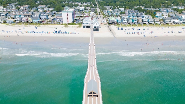 An aerial view of a coastal area with a long pier extending into the water, lined with beach houses and a beach with umbrellas and people.
