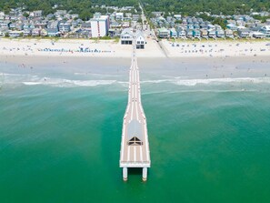 An aerial view of a coastal area with a long pier extending into the water, lined with beach houses and a beach with umbrellas and people. - Surfside Shamrock, 6 bed, 4.5 bath, easy walk to the beach or Surfside Pier! (Surfside Beach)