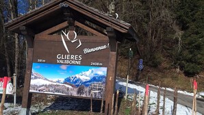 A wooden sign with a mountain view and distance markers, welcoming visitors to a ski resort.