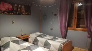 A bedroom with two beds, a wooden nightstand, a wall-mounted shelf with decorative items, and a window with curtains.