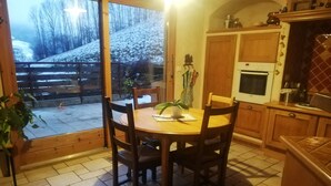 A kitchen with wooden cabinets, a dining table with chairs, a potted plant, and a view of a snowy landscape through a glass door.