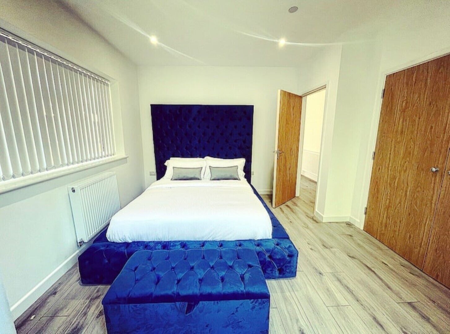 A bedroom with a large bed, a dark headboard, a wooden wardrobe, and a window with blinds.