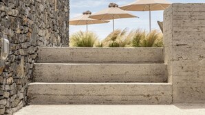 A set of concrete steps leading up to a stone wall, with outdoor umbrellas and grass in the background. - Korfi de Milo (Milos)