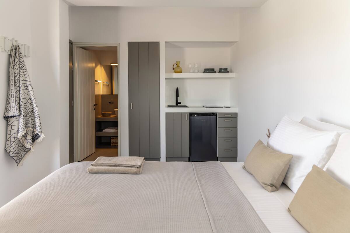 A bedroom with a bed, pillows, a bedside table, a wardrobe, and a small kitchenette area with a sink and storage.