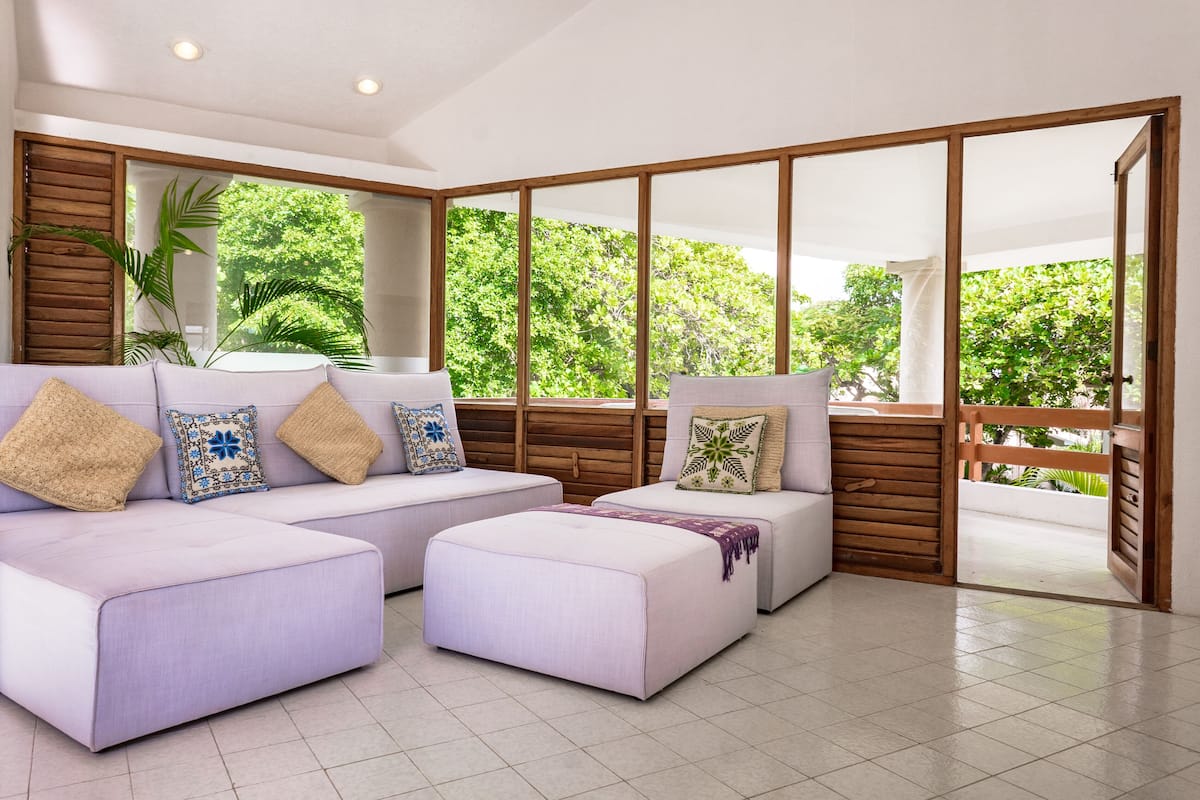 A modern living room with a sectional sofa, ottoman, and a large sliding glass door leading to an outdoor area with greenery.