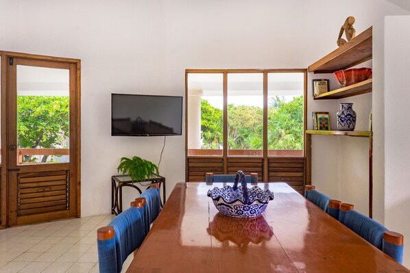 A dining area with a wooden table, blue cushioned chairs, a decorative blue and white bowl, a television on the wall, and a view of greenery through large windows.