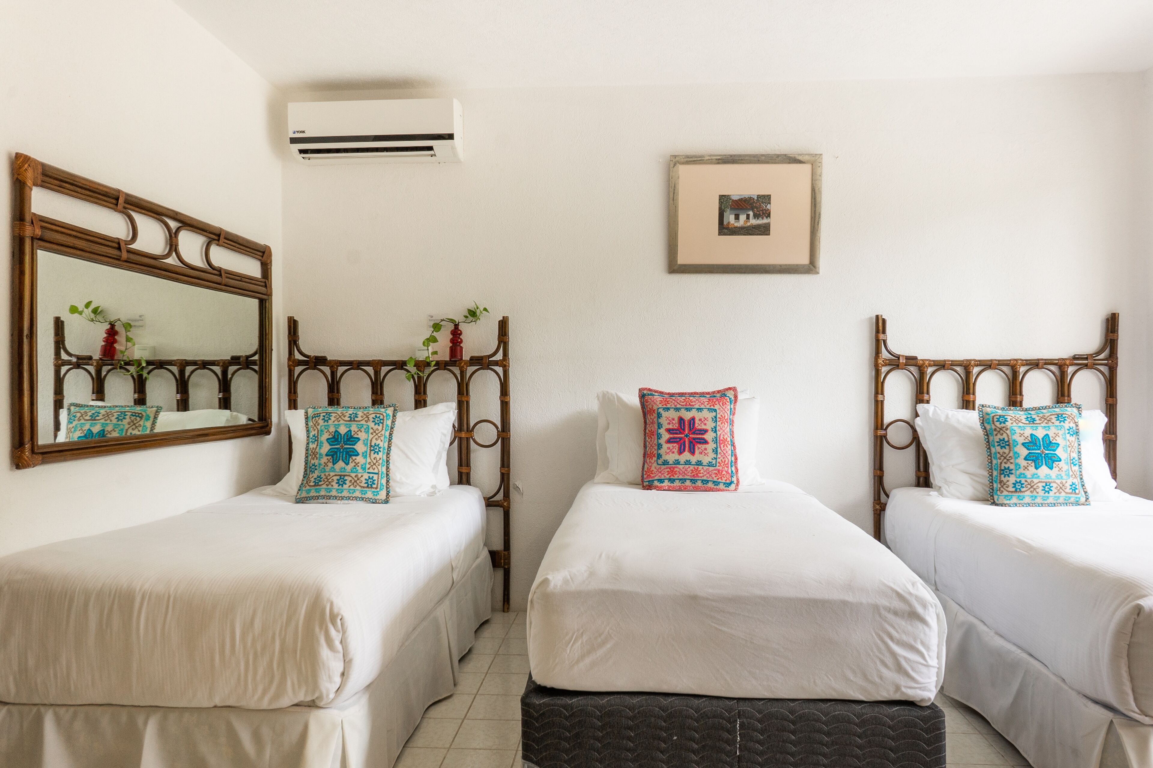 A hotel room with two beds, each with a headboard, white bedding, and decorative pillows. There is a wall-mounted air conditioner and a framed picture above the beds.