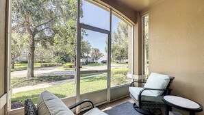 A sunroom with a view of a residential street, a cushioned chair, a small table, and a wicker bench.