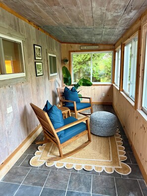 A wooden-paneled room with two wooden chairs with blue cushions, a round ottoman, and a large window with a view of greenery.