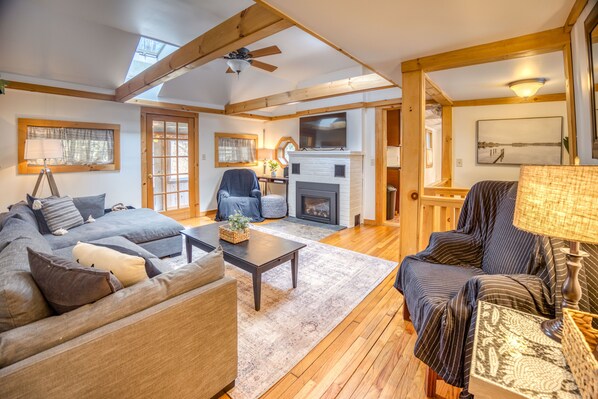 A cozy living room with a fireplace, wooden beams, and a ceiling fan.