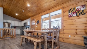 A rustic cabin interior with a wooden dining table, benches, and a kitchen area with white cabinets and a microwave.